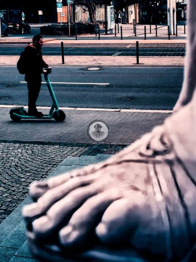Street One I 2023 WEB Jemand fährt auf einem Elektro-Roller an dem Fuss einer antiken Statue vorbei